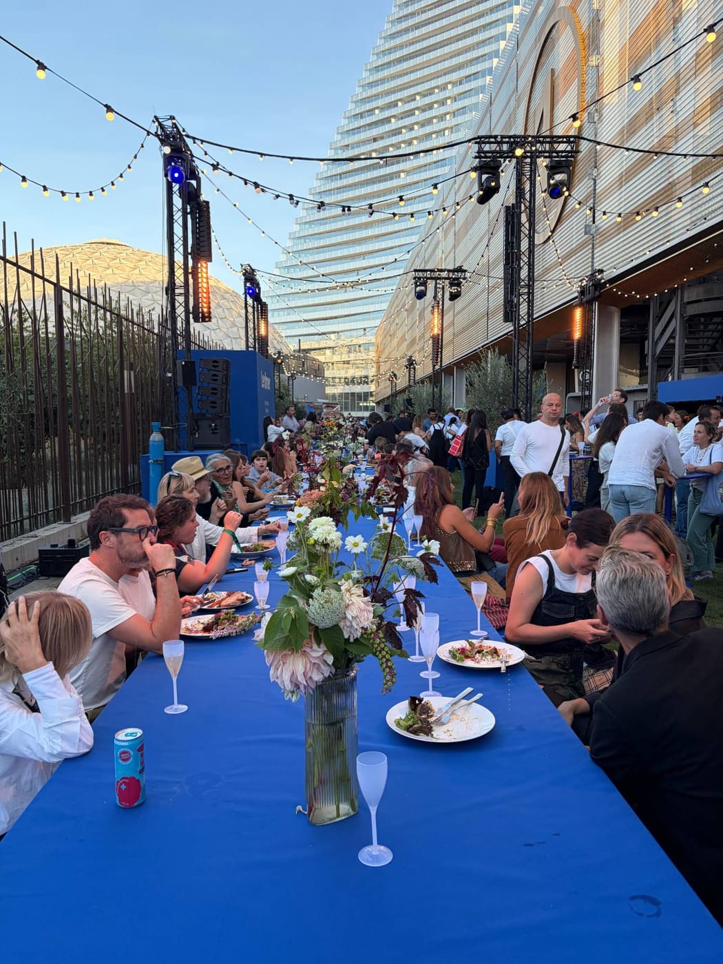 Guests at a long blue dinner table under string lights outside Hall 1, Paris expo Porte de Versailles, during Le Dîner at Who's Next, September 2026.